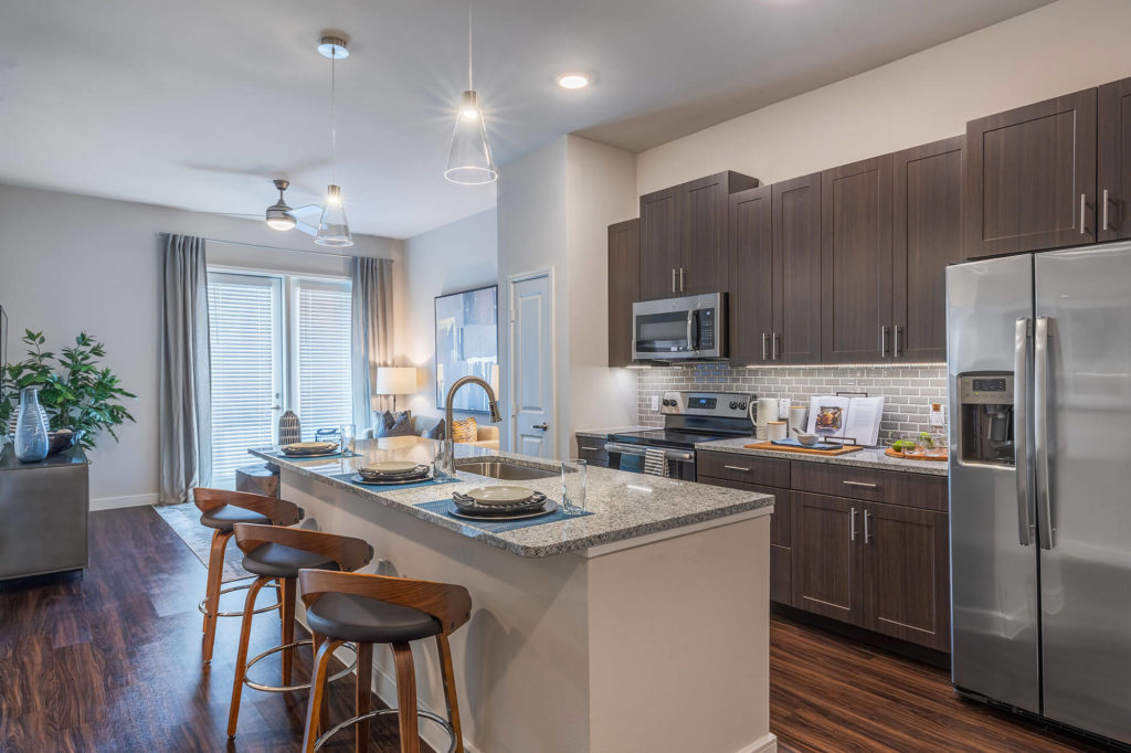 Open concept kitchen and living room with wood-style floors, granite counters, stainless-steel appliances, kitchen island with room for 3 seating, decorative pendant lighting, ceiling fan, and door to patio